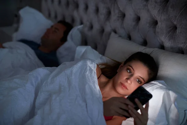 Mujer Acostada Cama Revisando Teléfono Móvil Mientras Hombre Duerme Junto —  Fotos de Stock