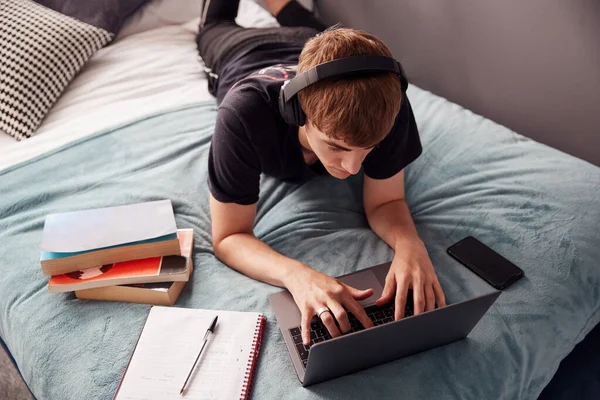 大学生头戴耳机躺在床上在笔记本电脑上工作的现象 — 图库照片