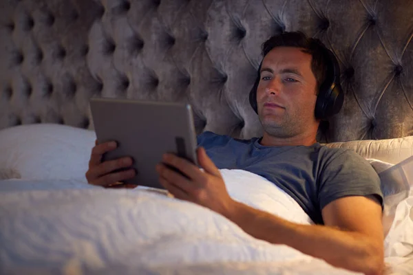 头戴无线耳机的男人在睡觉前躺在床上看数字表 — 图库照片