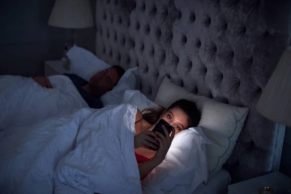 Kobieta Leżąca Łóżku Sprawdza Telefon Komórkowy Podczas Gdy Mężczyzna Śpi — Zdjęcie stockowe