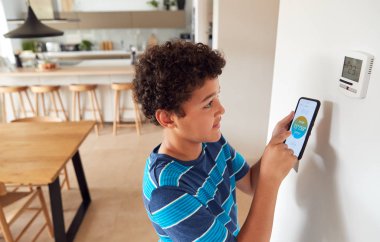 Boy Changes Temperature On Central Heating Thermostat Control Using Mobile Phone App clipart