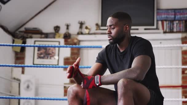 ボクシングリングの横に座って手に保護ラップを置くジムの男性ボクサー スローモーションで撮影 — ストック動画