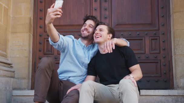 男性ゲイカップル座っています上の建物の階段の上に市内の通り取りますセルフィーオン携帯電話 ショットでスローモーション — ストック動画