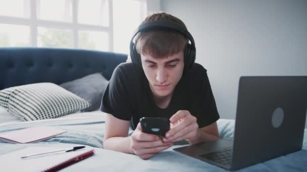 男大学生头戴耳机躺在床上 在笔记本电脑上工作 用手机射击 动作缓慢 — 图库视频影像