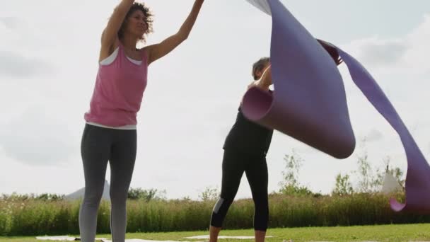 两名成熟的女性朋友在室外瑜伽静修会上在草地上展开运动垫 动作缓慢 — 图库视频影像