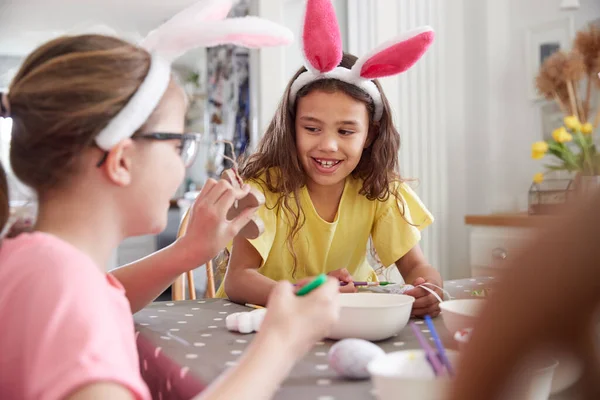 三个女孩穿着兔子耳朵坐在桌旁装饰彩蛋回家过复活节 — 图库照片