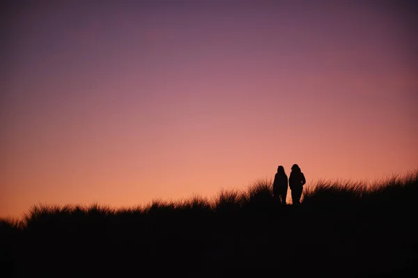 两个女性朋友与夕阳西下走过沙丘的轮廓 — 图库照片
