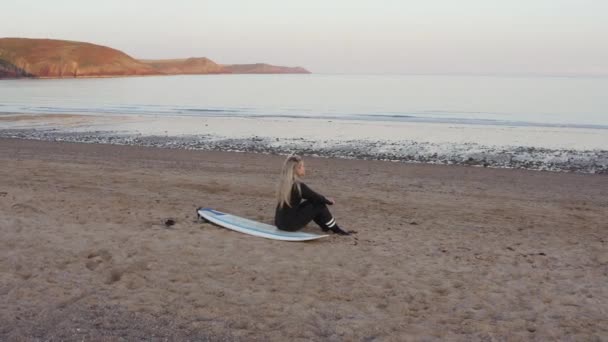ドローンショットの女性はウェットスーツを着てサーフボードに座って 日没時に海を見下ろす スローモーションで撮影 — ストック動画