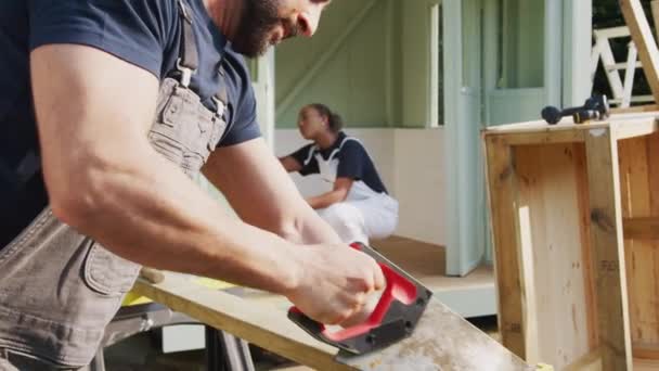 Snickare Sågar Trä Förgrunden Som Han Och Lärling Bygga Sommarhus — Stockvideo