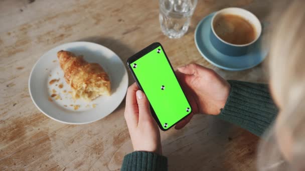 坐在桌边喝咖啡和吃羊角面包的女人在手机上翻看绿色屏幕的照片 动作缓慢 — 图库视频影像