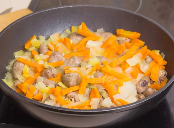 Los corazones de pollo fritos con verduras, zanahorias, cebolla, verduras en una sartén negra, cerraron la tapa de vidrio. Primer plano de fondo. Preparar comidas en la cocina . — Foto de Stock