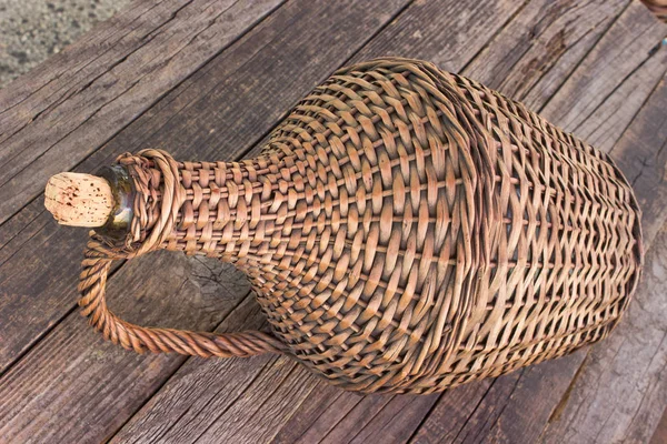 Old demijohn wicker wrapped glass bottle on wooden table — Stock Photo, Image