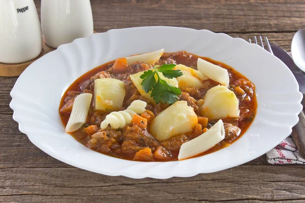 Gulasch-Eintopf mit Kartoffeln und Nudeln im Teller auf Holztisch — Stockfoto