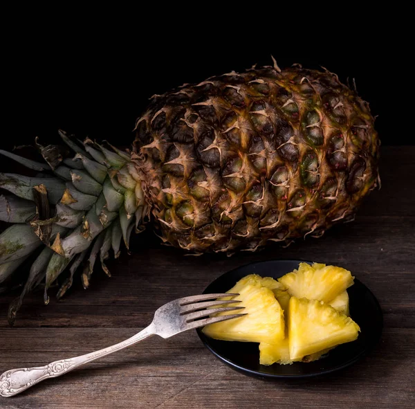 Sliced pineapple on old rustic desk with knife and with whole pineapple behind. Black background.