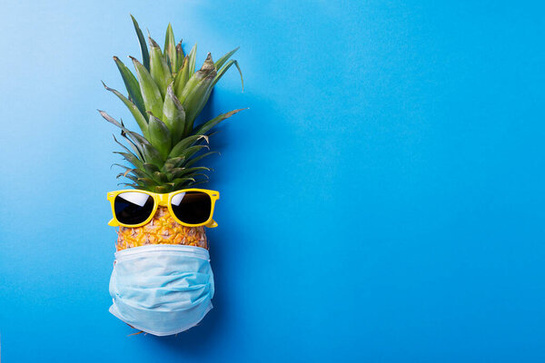 Concept of summer vacation 2020. Pine apple with a medical mask and sun glasses on the blue background. Top view image