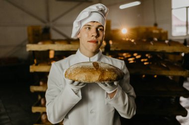 Bir fırıncı elinde taze ekmek tepsisiyle fırının arka planındaki raflara karşı taze ekmek tutar. Sanayi ekmek üretimi