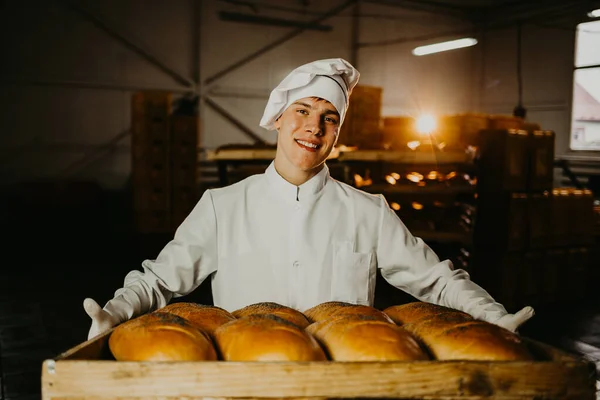 A baker holds a tray with fresh hot bread in his hands against the background of shelves with fresh bread in a bakery. Industrial bread production