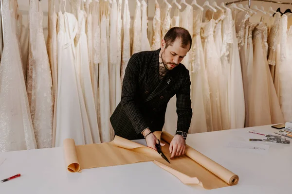 Young serious tailor with beard looking on drawing near wooden table with threads in amazing atelier with antique furniture and mannequin