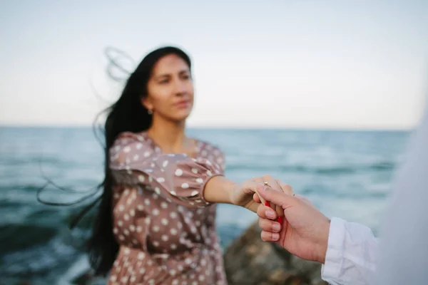 ビーチで手と心を提供しています 男は指輪を与え 女の子に結婚するように頼むと 彼女は満足しており 彼女の手に彼女の顔を保持します 崖の上の海と岩場のビーチの周り — ストック写真