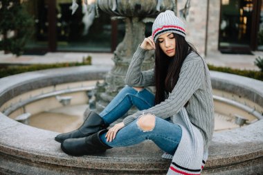 Şapka ve eşarp takmış genç bir kız açık havuzun yanında oturuyor..