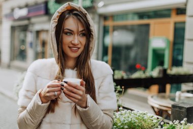 Kış kenti caddesinde elinde bir fincan çay ya da kahveyle şık gülümseyen bir kadın. Koyu mavi ceketli, kürklü gri şapkalı gülümseyen kız parkta yürüyor..