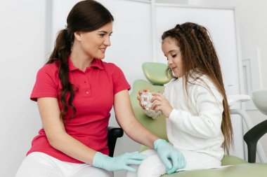 Küçük kız dişçide sandalyede oturuyor ve yanında mutlu bir doktor var.