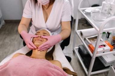 Kadın kozmetikçi, ofisteki masada yatan hastanın yüzünü bantlıyor.