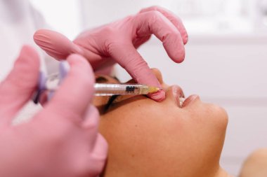 Kadın dudaklarına botoks yapan kozmetik uzmanının ellerine yakın çekim. Elinde şırınga var. Genç ve güzel bayan keyifle prosedür uygulanıyor.