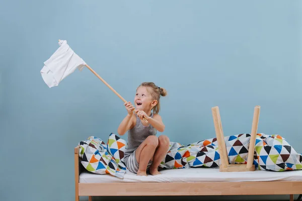 Menino está sentado na cama desfeita, acenando bandeira branca sobre paredes azuis — Fotografia de Stock