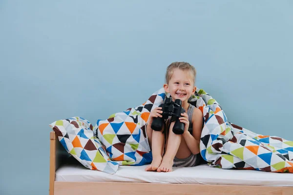 Little boy is playing in bed holding binoculars in hands over blue walls — Stock Photo, Image