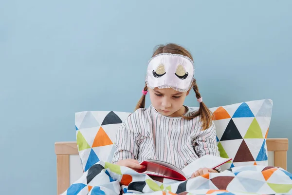 Klein meisje is het lezen in bed in een pyjama voordat je gaat slapen over blauw — Stockfoto