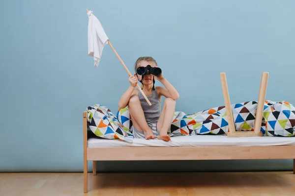 Kleine jongen zit op onopgemaakt bed met witte vlag, kijkend door een verrekijker — Stockfoto