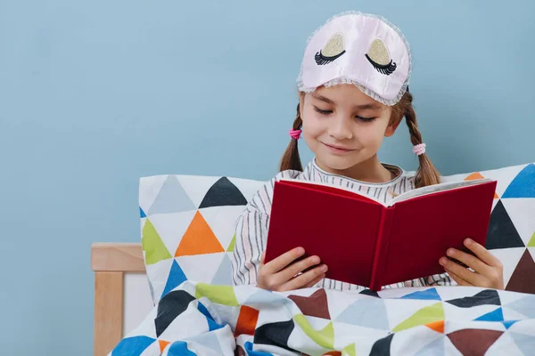 Ijverig klein meisje lezen in bed in pyjama voor gaan slapen over blauw — Stockfoto