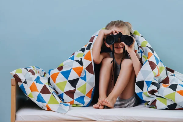 Kleine jongen zit op onopgemaakt bed, kijkend door een verrekijker over blauwe muren — Stockfoto