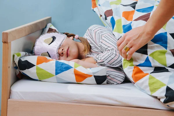 Mother tucks her little daughter in bed. Girl wearing sleeping mask. — Stock Photo, Image