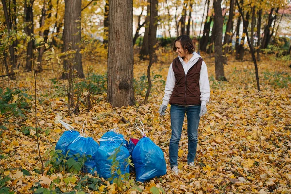 Kvinnan står bredvid en hög med blå plastpåsar i en skog på hösten. — Stockfoto