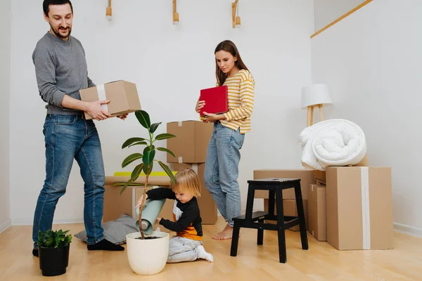 A família mudou-se para um novo apartamento. Planta de rega infantil enquanto os pais desempacotam . — Fotografia de Stock