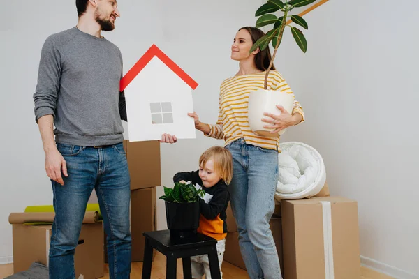 A família mudou-se para um novo apartamento. Planta de rega infantil enquanto os pais desempacotam . — Fotografia de Stock