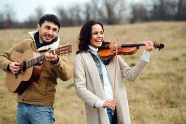 Sıcak giyimli müzisyenler kırsalda çalarken birlikte gülerler.