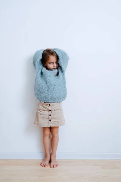 Egy kis mezítlábas lány kék pulóverben, aki a falnak támaszkodik otthon. — Stock Fotó