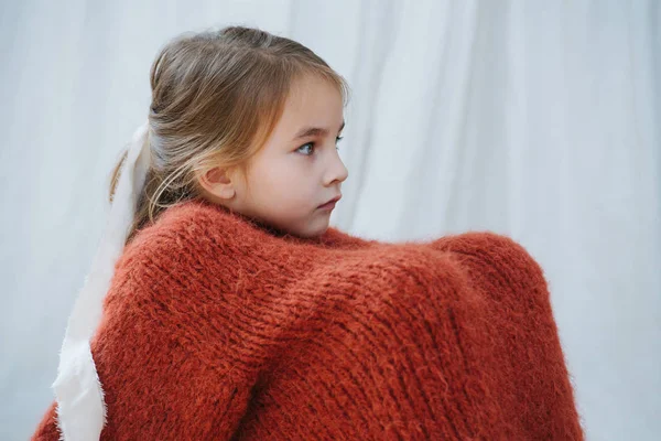 Niña sentada con las rodillas envuelta en un suéter de punto naranja en casa —  Fotos de Stock