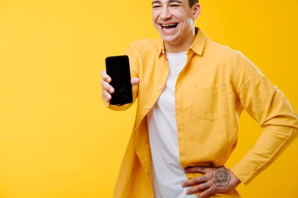 Lange lachende jongeman in geel shirt met foto op zijn telefoon over geel — Stockfoto