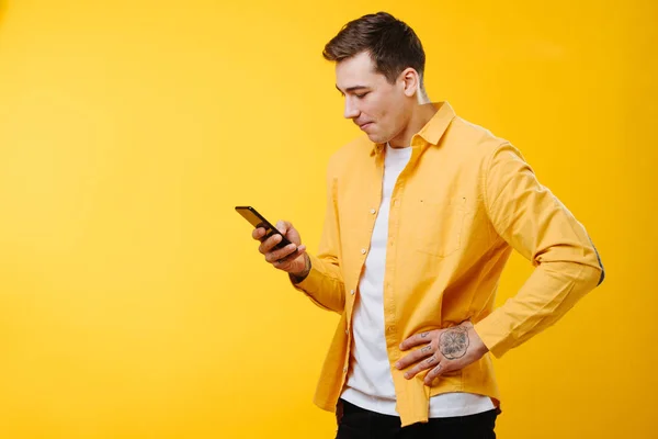 Knappe brutale jongeman in een geel shirt die zijn telefoon over geel checkt. — Stockfoto