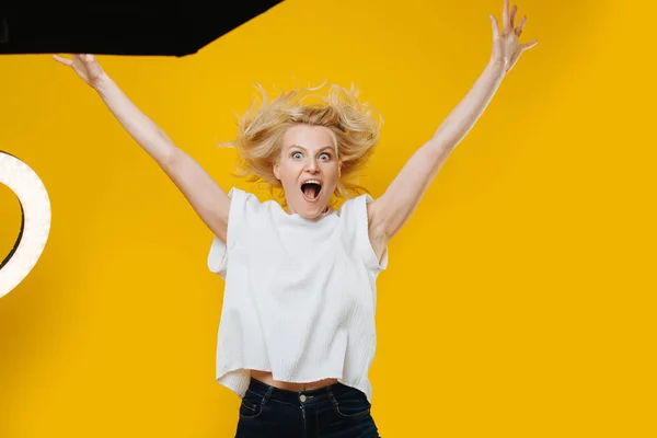 Игривая блондинка, прыгающая с поднятыми руками над жёлтым — стоковое фото