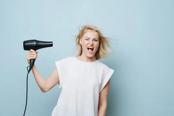 遊び心ブロンド女性打撃自分の顔にAヘアドライヤーとともに青 — ストック写真