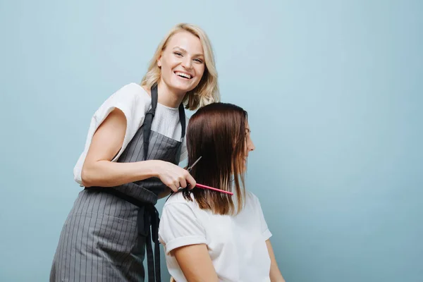 Alegre loira fêmea cabeleireiro pentear o cabelo do cliente sobre azul fundo . — Fotografia de Stock