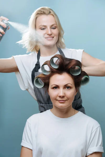 Mulher estilista pulverizando cabelo castanho do cliente em rolos sobre azul — Fotografia de Stock