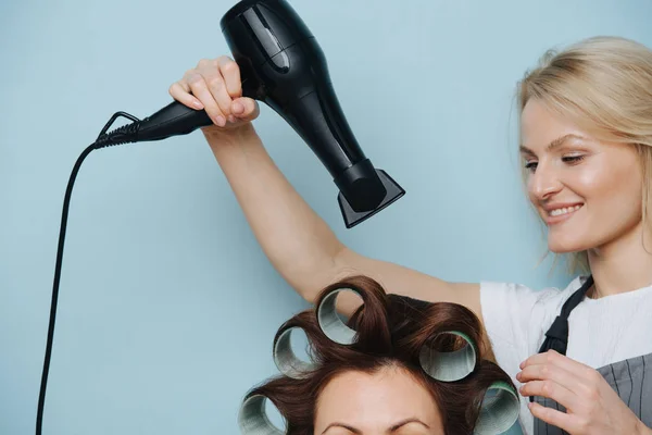 Charmoso cabeleireiro senhora secagem cabelo castanho do cliente em rolos sobre azul — Fotografia de Stock