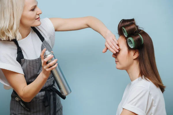 Loira estilista feminino pulverização cabelo castanho do cliente em rolos sobre azul — Fotografia de Stock