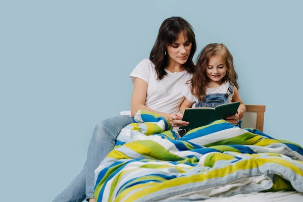 Mom reads book to her daughter sitting on the bed.